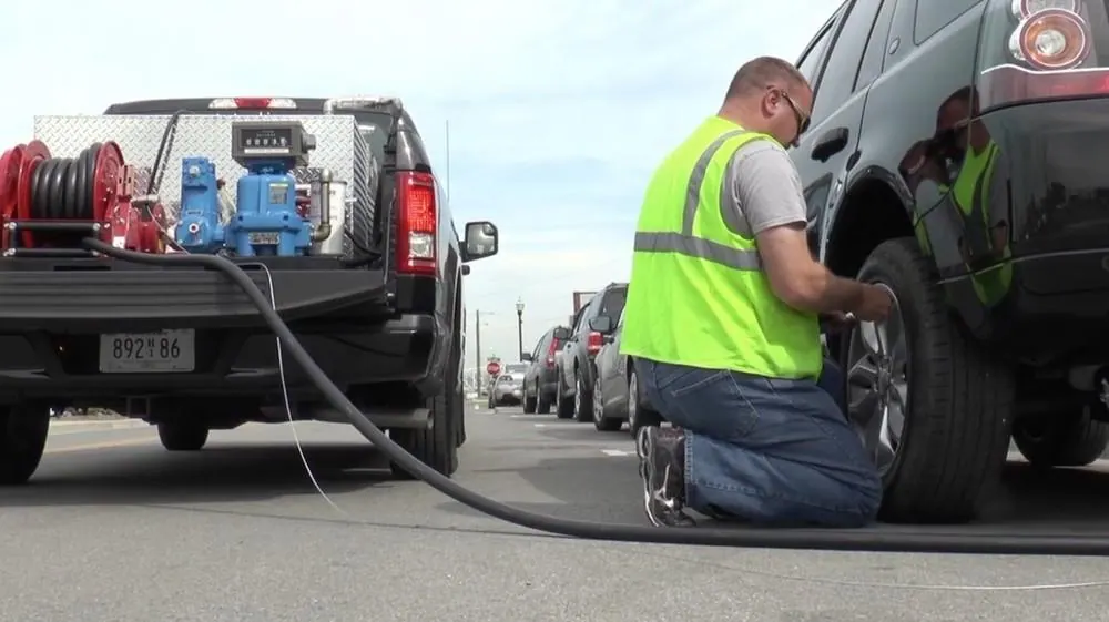 Pickup truck fuel delivery rig with a red hose reel, blue pump and mechanical meter register in the bed, its hose running to a worker in a safety vest kneeling beside a parked SUV