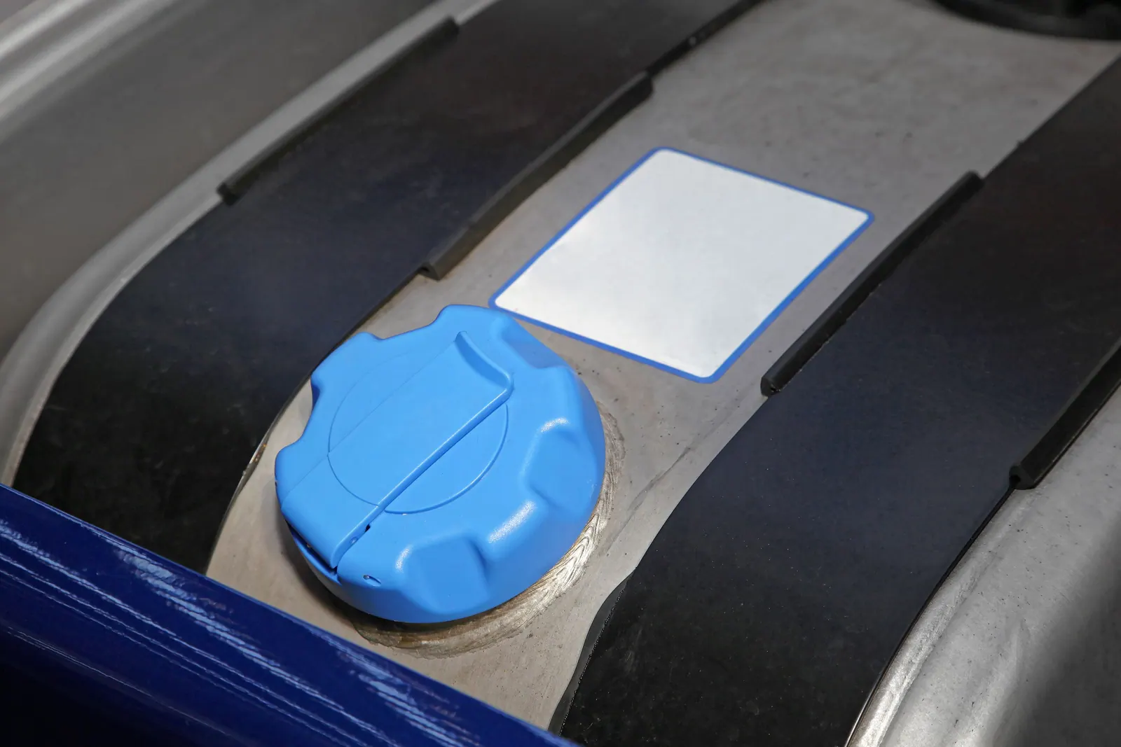 Blue diesel exhaust fluid filler cap on a truck's DEF tank, next to a blank blue-bordered label