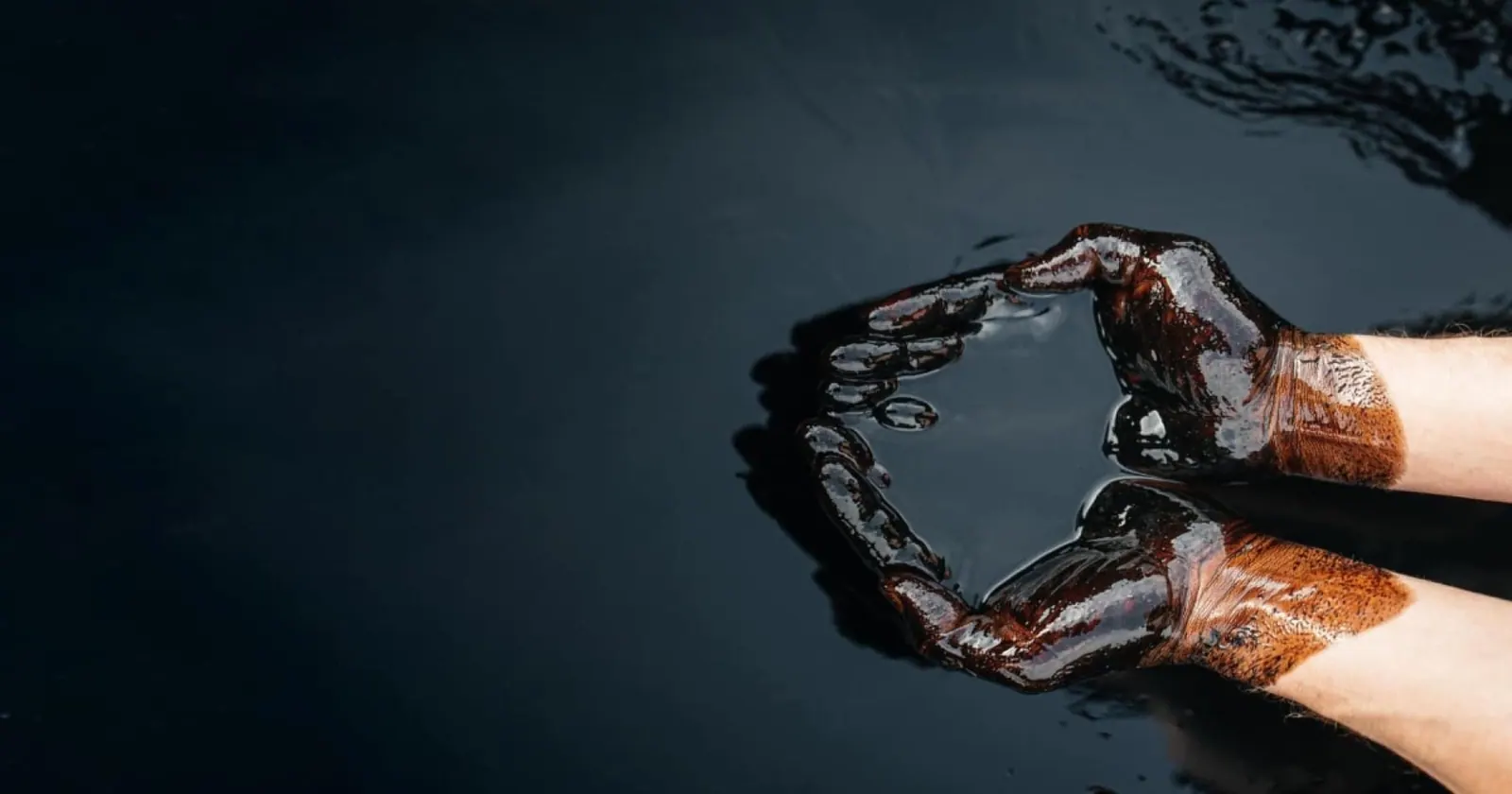 Two hands coated in crude oil, cupped above a dark pool of crude